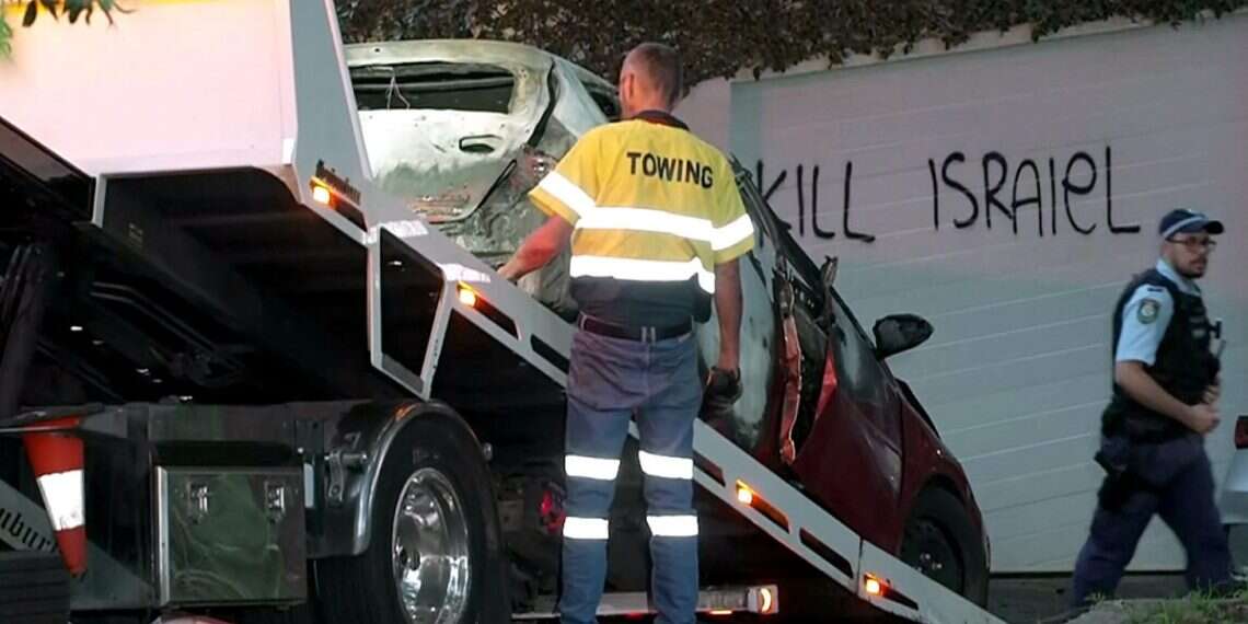 This frame grab taken from video footage provided by the ABC on Dec. 11, 2024 shows a police officer walking past a torched car being removed in front of anti-Israel graffiti written on a wall in the Sydney suburb of Woollahra (Handout/Australian Broadcasting Corporation/ AFP) Sydney, Melbourne Jewish communities targeted with arson, graffiti
